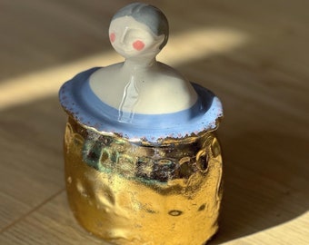 May include: A ceramic jar with a gold finish and a lid shaped like a woman's head. The lid is painted with a blue band and the woman's face has pink cheeks.
