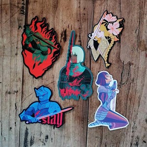 May include: A collection of five die-cut stickers featuring stylized illustrations of armored figures with swords. The stickers showcase a variety of color palettes, including red, blue, green, and gold, with some designs incorporating flames and floral elements.