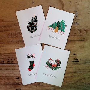 Könnte beinhalten: Vier Weihnachtskarten mit festlichen Illustrationen. Eine Karte zeigt eine schwarze Katze mit Lichterketten, eine andere einen Weihnachtsbaum mit einer Katze, einen Strumpf und eine Katze in einer Geschenkbox. Jede Karte hat einen anderen Feiertagsgruß.