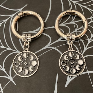 May include: Two silver keyrings with black and white moon phase charms. The charms are round and feature a detailed design of the moon in different phases.
