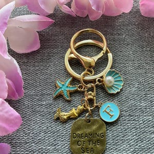 May include: Gold keyring with a blue and white seashell charm, a blue starfish charm, a mermaid charm, and a square charm with the text "Dreaming of the Sea".