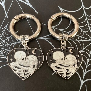 May include: Two silver keyrings with black heart-shaped charms. Each charm features a white skeleton couple kissing with a crescent moon and stars on a black background.
