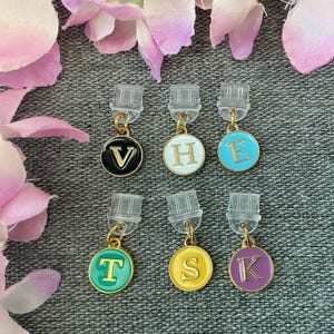 May include: Six initial earrings with gold-tone metal detailing. Each earring displays a different letter: V, H, E, T, S, and K, set against a black, white, turquoise, green, yellow, and purple background. The earrings are presented on a grey textured surface.