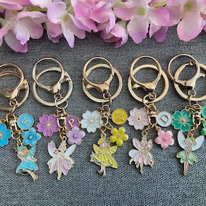 May include: Five gold keyrings with colourful enamel charms. Each charm features a different letter, a flower, and a fairy design. The charms are attached to a gold split ring.