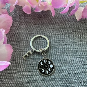 May include: A silver keyring with a charm featuring a black and white moon phases design. The charm is attached to a silver ring with a letter "E" charm.