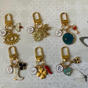 May include: Six gold-tone keyrings with assorted charms. Charms include an eye, a cloud, a leaf, a leopard, a wine glass, a moon, a clover, a boot, a chilli pepper, and a heart with an eye. Each keyring has a gold clasp and a white initial charm.