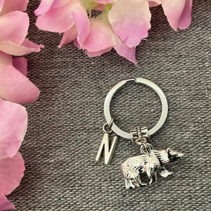 May include: A silver-toned metal keyring featuring a circular split ring, a dangling letter N charm, and a detailed bear-shaped charm. The bear charm has a textured finish that highlights its form, and the letter N is a simple block design.
