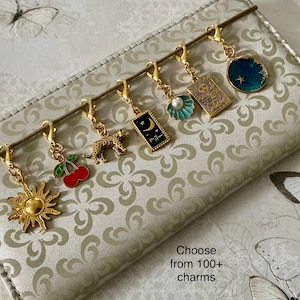 May include: A set of six gold-toned metal charms with lobster clasps, including a sun, red cherries, a leopard, a black rectangular charm with a moon, a teal seashell with a pearl, and a circular blue charm with a star, plus text: Choose from 100+ charms.