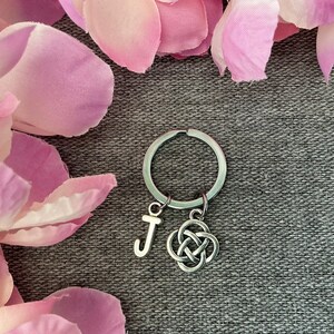 May include: A silver-toned metal keyring featuring a circular split ring, a letter J charm, and a separate intricate Celtic knot charm. The keyring is constructed from polished metal components and displays a clean, metallic finish throughout.