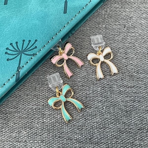 May include: Three bow-shaped stud earrings in pink, white, and teal with gold-coloured outlines. The earrings are displayed on a grey surface next to a teal notebook.