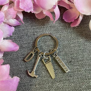 May include: A silver-toned metal keyring featuring four charms attached to a circular ring: a letter D, a hammer, a hand saw, and a ruler. The charms are crafted from a metallic material with a smooth finish, showcasing detailed tool shapes.
