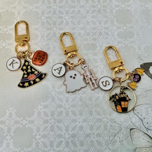 May include: Three gold-toned metal keychains featuring Halloween-themed charms. One has a witch hat, a pumpkin, and the letter K. Another has a ghost, a skeleton, and the letter A. The third has a haunted house, a bat, and the letter S.