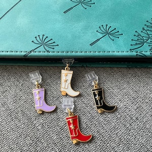 May include: Four colourful cowboy boot earrings. The earrings are in the colours of purple, white, black, and red. Each boot has a gold star and gold accents. The earrings are hanging from clear earring backs.