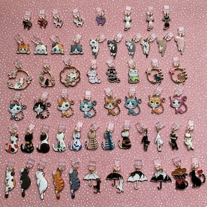 May include: A selection of cat-themed charms in various colours and designs. The charms are attached to clear clips, and include cats with floral accents, crowns, and umbrellas. The background is a pink surface with small white dots.