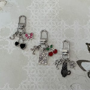 May include: Three silver keyrings with decorative charms. One features black cherries, a pink bow, and the letter "S". Another has red cherries, a tarot card, and the letter "R". The last has a black cat, a four-leaf clover, and the letter "R".