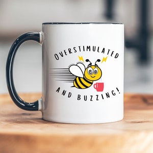 May include: White ceramic mug with a black handle and rim. The mug has a cartoon bee with a cup of coffee and the text "Overstimulated and Buzzing!" printed on it.