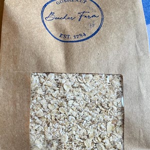 May include: A brown paper bag with a clear window displaying rolled oats. The bag has a blue circular stamp that reads "Goshen, CT Beecher Farm EST. 1784". The oats are light brown and fill the window.
