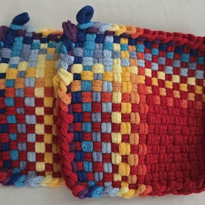 Peut inclure: Deux maniques tissées colorées avec un schéma de couleurs rouge, orange, jaune, bleu et violet. Les maniques sont de forme carrée et ont une texture tissée.