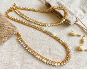 May include: A gold-toned chain necklace featuring a row of clear, sparkling rhinestones embedded along the length of the metal links. The necklace is arranged in a curved shape, showcasing the reflective quality of the stones and the metallic finish.