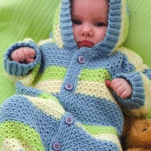 May include: A baby wearing a handmade crochet jumpsuit with a hood. The jumpsuit features horizontal stripes of blue, green, and cream colors. It has a button-up front and drawstring ankles. The baby is lying on a green surface.