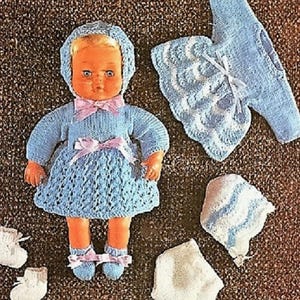 May include: A vintage doll wearing a light blue knitted dress, bonnet, and booties. The dress has an openwork skirt and pink bows. A matching knitted jacket and hat are also visible. The ensemble evokes a classic, handmade aesthetic, with a focus on detail.