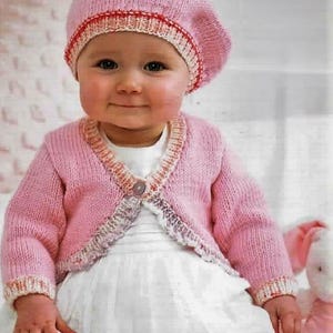 May include: A baby wearing a pink knitted beret and matching cardigan. The beret has a red and white trim, and the cardigan has a single button closure. The baby is also wearing a white dress. The overall look is soft and cozy.