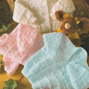 May include: Three knitted baby cardigans in pastel shades of white, pink, and light blue. The garments feature a textured knit design and long sleeves. A teddy bear with a yellow bow is partially visible.