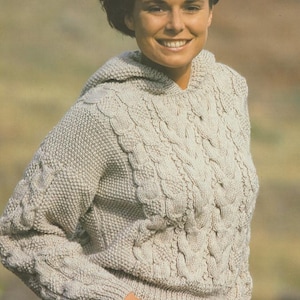 Puede incluir: Un suéter con capucha de color beige claro con intrincados detalles de punto de cable. El suéter presenta un patrón texturizado en el pecho y las mangas, con dobladillo y puños acanalados. La capucha es visible y el suéter se usa con jeans azules.