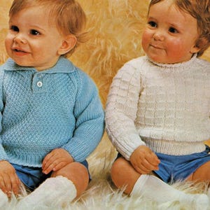 May include: Two babies wearing knitted sweaters and shorts. One baby is wearing a light blue sweater with a collared neckline and a single button. The other baby is wearing a white sweater with a crew neck. Both babies are wearing blue shorts and white socks.
