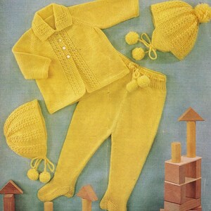 May include: A complete yellow knitted baby outfit, including a collared cardigan with button closure, matching pants, and two knitted hats with pom-poms. The set is displayed on a light blue background, with wooden blocks in the foreground.