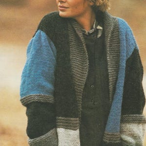 Puede incluir: Un cárdigan de punto con bloques de color en tonos azul, negro, gris y blanco. El suéter de manga larga tiene cuello chal y puños acanalados. El modelo lleva una camisa oscura debajo del cárdigan.