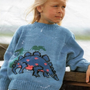 Peut inclure: Un pull bleu clair tricoté avec un grand dinosaure bleu à pois roses et des accents verts. Le motif du dinosaure est centré sur le devant. Le pull a un col et des poignets côtelés.