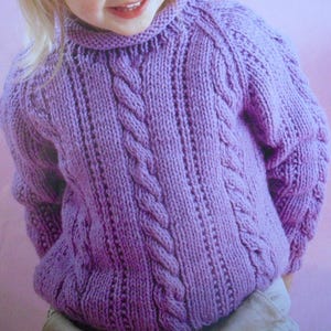 May include: A lavender-colored, hand-knitted sweater with a cable knit design on the front. The sweater has a ribbed collar and cuffs. The sleeves are long and the overall style is cozy and warm. The sweater is made of a soft, textured yarn.