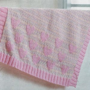 May include: A soft, pink and white knitted blanket with a repeating pattern of raised pink hearts. The blanket has a ribbed pink border, creating a cozy and textured appearance. This blanket is perfect for a baby.