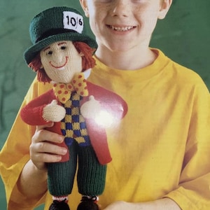 May include: A knitted doll dressed as the Mad Hatter, featuring a green hat with "10/6" on it, a red coat, and a checkered vest. The doll has orange hair, a yellow bow tie, and green pants. The doll is being held by a person wearing a yellow shirt.