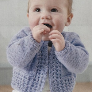 May include: A light purple baby cardigan with a textured pattern on the front and sleeves. The cardigan is open in the front and has a ribbed hem and cuffs. The baby is wearing a white onesie underneath.