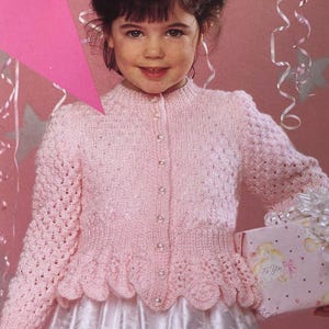 May include: A light pink knitted cardigan with pearl buttons and a scalloped hem. The long-sleeved sweater has a textured pattern and is worn over a white dress. The image shows a young girl holding a gift box.
