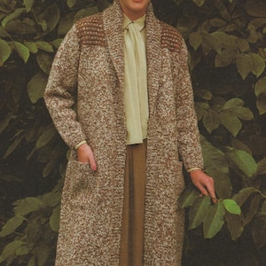May include: A long, brown and white speckled knit coat with a shawl collar and pockets. The coat is worn over a light-colored blouse and skirt. The woman is smiling in front of a backdrop of green leaves.