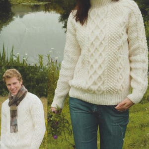 May include: Cream-colored cable-knit sweater with a turtleneck. The sweater features a textured diamond pattern on the front and sleeves. The image shows a person wearing the sweater with blue jeans. A second person is wearing a similar sweater.