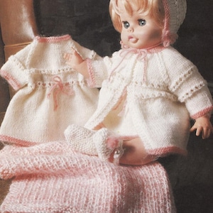 Può includere: Una bambola vintage che indossa un completo in maglia bianco con rifiniture rosa, tra cui cappotto, cappello e scarpine. Sono presenti anche un vestito abbinato e una coperta in maglia rosa. La bambola ha i capelli biondi e gli occhi azzurri.