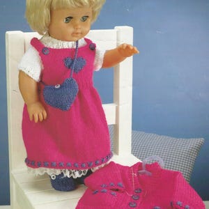 Può includere: Bambola con un vestito rosa lavorato a maglia con una tasca blu a forma di cuore e scarpe blu abbinate. Una giacca rosa lavorata a maglia con dettagli blu è vicina. La bambola ha i capelli biondi ed è seduta su una sedia bianca.