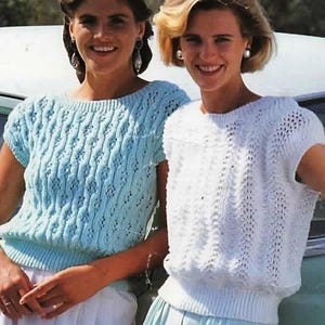 May include: Two women wearing knitted tops. One top is light blue, the other is white. Both tops have a textured, open-weave design and short sleeves. The women are smiling, and the tops appear to be part of a vintage fashion collection.