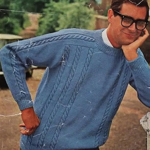 Puede incluir: Un suéter de punto trenzado azul claro con cuello redondo. El suéter presenta un diseño texturizado con patrones de cable verticales y horizontales. Las mangas y los hombros tienen un diseño trenzado. La persona de la imagen lleva gafas negras y una camiseta interior blanca.