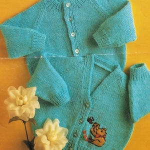 May include: Two turquoise knitted baby cardigans. One cardigan has a button-up front and long sleeves. The second cardigan has a button-up front, long sleeves, and a Winnie the Pooh embroidery design. The cardigans are displayed on a yellow background.
