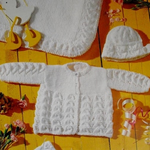 Op de afbeelding: Een set witte gebreide babykleding, waaronder een deken, muts, vest en babyslofjes. Het vest heeft een knoopsluiting en textuurpatronen. De items worden gepresenteerd op een gele ondergrond met decoratieve accenten.