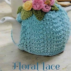 Puede incluir: Un calentador de té de punto azul claro con un diseño floral, rematado con flores de hilo rosa, amarillo y verde. El calentador de té está sobre una tetera blanca, con una taza de té y galletas en el fondo. El texto "Floral Lace Tea Cosy" está en la parte inferior.