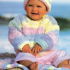 May include: A baby in a pastel-striped sweater and hat, with pink trousers. The sweater features horizontal stripes of yellow, pink, blue, and green. The baby is sitting on a colourful blanket, with bare feet visible.