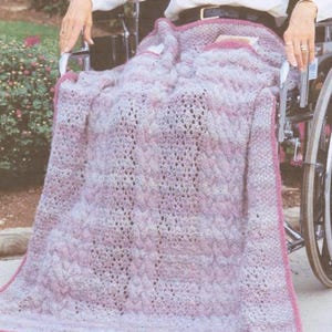 May include: A soft, knitted throw blanket featuring a variegated color palette of light pink, lavender, and white. The blanket displays a textured pattern with vertical cable knit columns interspersed with delicate, openwork lace sections.