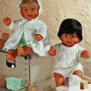 May include: Two baby dolls wearing handmade knitted outfits. One doll is wearing a white hooded coat over a green dress, the other is in a white top and shorts set. Both dolls have dark eyes and are sitting on a metal box.