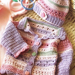 May include: A handmade crochet baby set in pastel colors, including a hat and cardigan. The cardigan has a scalloped edge, button closure, and long sleeves. The hat features a small flower accent. The set is made with pink, purple, and green yarn.
